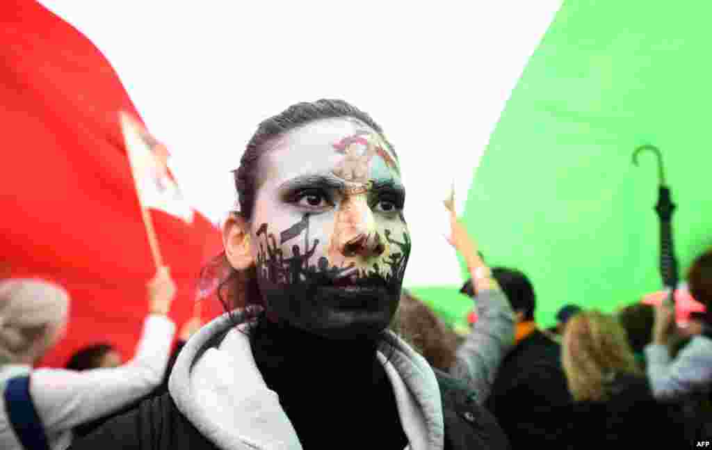 A protester wearing face-paint depicting France's iconic "Marianne" leading an uprising, stands under a giant Iranian flag as people gather in support of Iranian woman Mahsa Amini during a protest on Place de la Republique in Paris, following her death in custody.