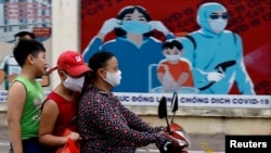 Người dân lái xe ngang qua một biểu ngữ cổ động phòng chống virus corona ở Hà Nội hôm 31/7/2020. Chính phủ Việt Nam đang đàm phán mua vaccine chống COVID-19 từ Mỹ và 3 nước khác trong khi tiếp tục thủ nghiệm lâm sàng để sản xuất vaccine nội địa.