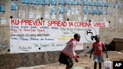 Vijana bawili wanauza njugu mbele ya tangazo linalowashauri watu kuchukua tahadhari dhidi kupata maambukizi mapya ya virusi vya corona, katika eneo la Kibera ambalo ni makazi ya watu yasiorasimishwa, Nairobi, Kenya, Alhamisi, Aprili 2, 2020.